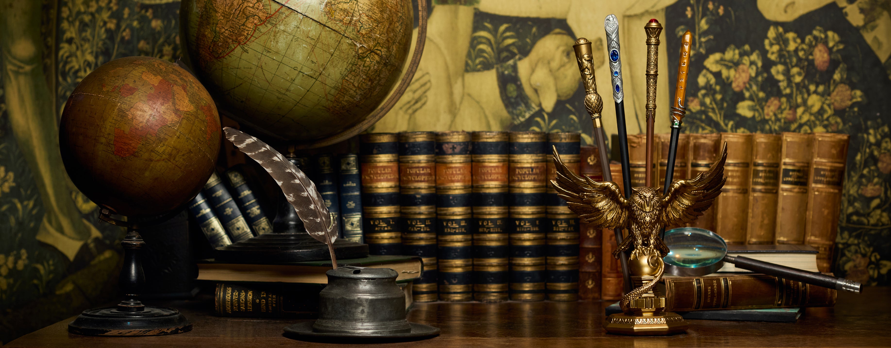 A vintage study still life featuring two antique globes, leather-bound books, a feather quill in a metal ink bottle, a gold owl wand stand, and a magnifying glass on a wooden surface against a patterned tapestry.