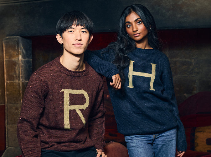 Two models wearing knit sweaters with H and R letters, posed in front of a dark fireplace. The model on the left wears a burgundy sweater with a gold 'R', and the model on the right wears a navy blue sweater with a gold 'H'.