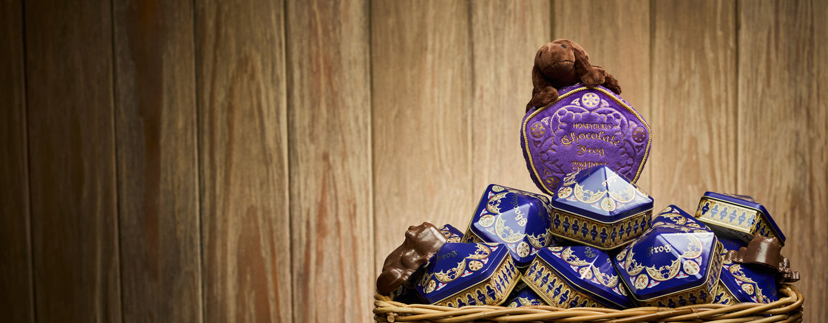 A collection of ornate blue and purple Chocolate Frog boxes with gold detailing, arranged in a wicker basket against a rustic wooden plank background.