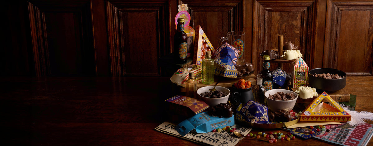 Darkly lit table with various confectionery items with bottles, bowls, and decorative objects against a wooden wall.