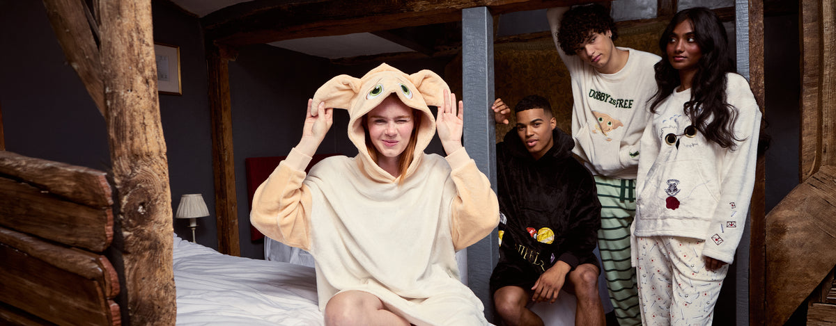 Four models wearing cozy loungewear and pajamas in a rustic bedroom setting with wooden beams. The front model wears a cream-colored Dobby hooded sweatshirt, while the others wear matching sweatshirts and pajama sets in black, green-and-white striped, and cream with Harry Potter creature prints.