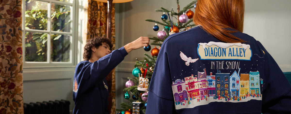 A person wearing a navy blue sweatshirt with a colorful 'Diagon Alley in the Snow' graphic on the back, standing indoors near a decorated Christmas tree while another person in a matching sweatshirt hangs ornaments.
