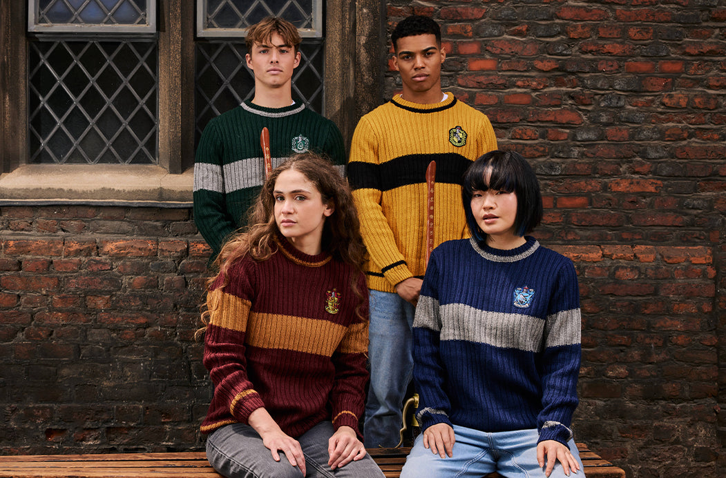 Four young adults wearing Quidditch-themed striped knit sweaters pose together in front of a rustic brick wall. The sweaters feature the Gryffindor, Slytherin, Hufflepuff, and Ravenclaw crests and are styled with wooden wand props.