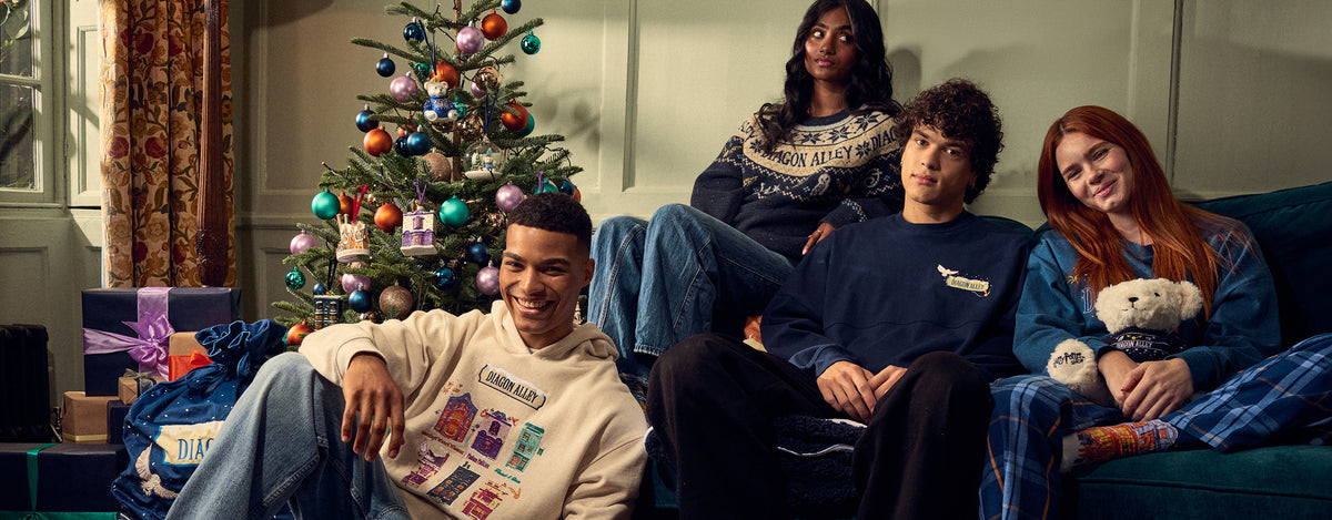 Four people sitting together on a couch in a cozy living room setting, with a decorated Christmas tree and wrapped gifts in the background. They are wearing matching navy blue and cream knit sweaters and pajama sets.