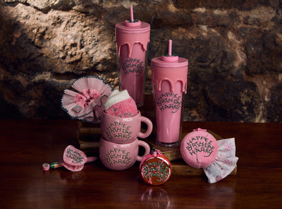 Pink tumblers and mugs with decorative elements on a wooden surface against a stone wall background.