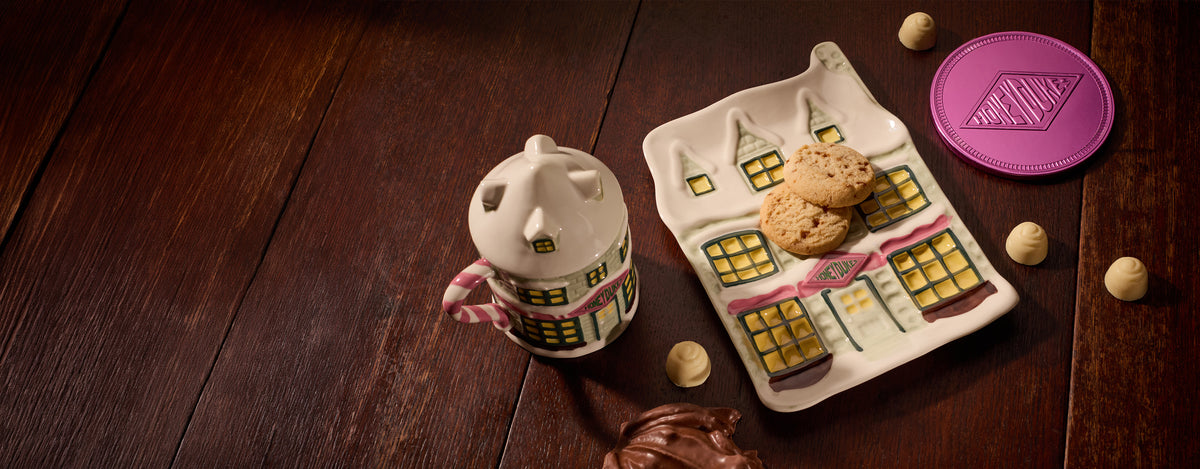 A ceramic mug shaped like Honeydukes with a pink and white handle and yellow window details sits beside a matching house-shaped plate holding two cookies, on a dark wooden tabletop with a few chocolates scattered around.