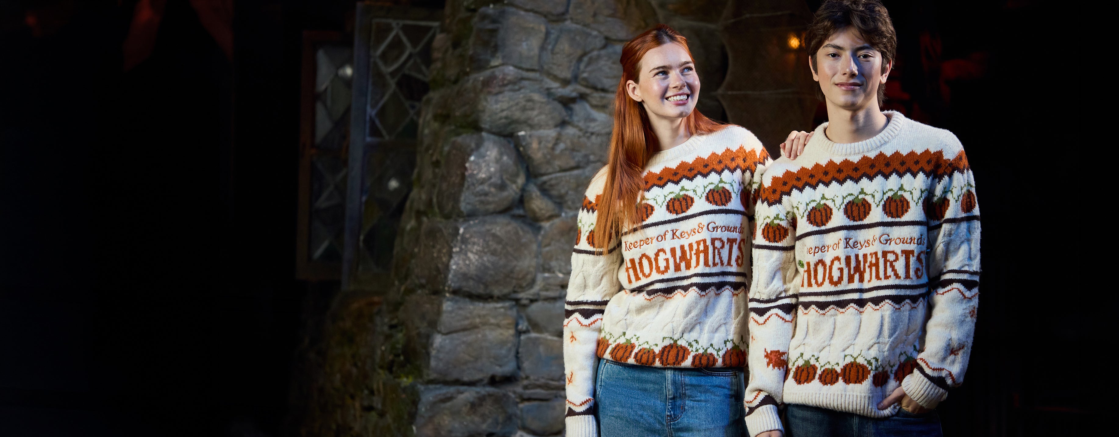 Two young adults wearing matching Hogwarts-themed knit sweaters, standing in front of a stone wall at night.
