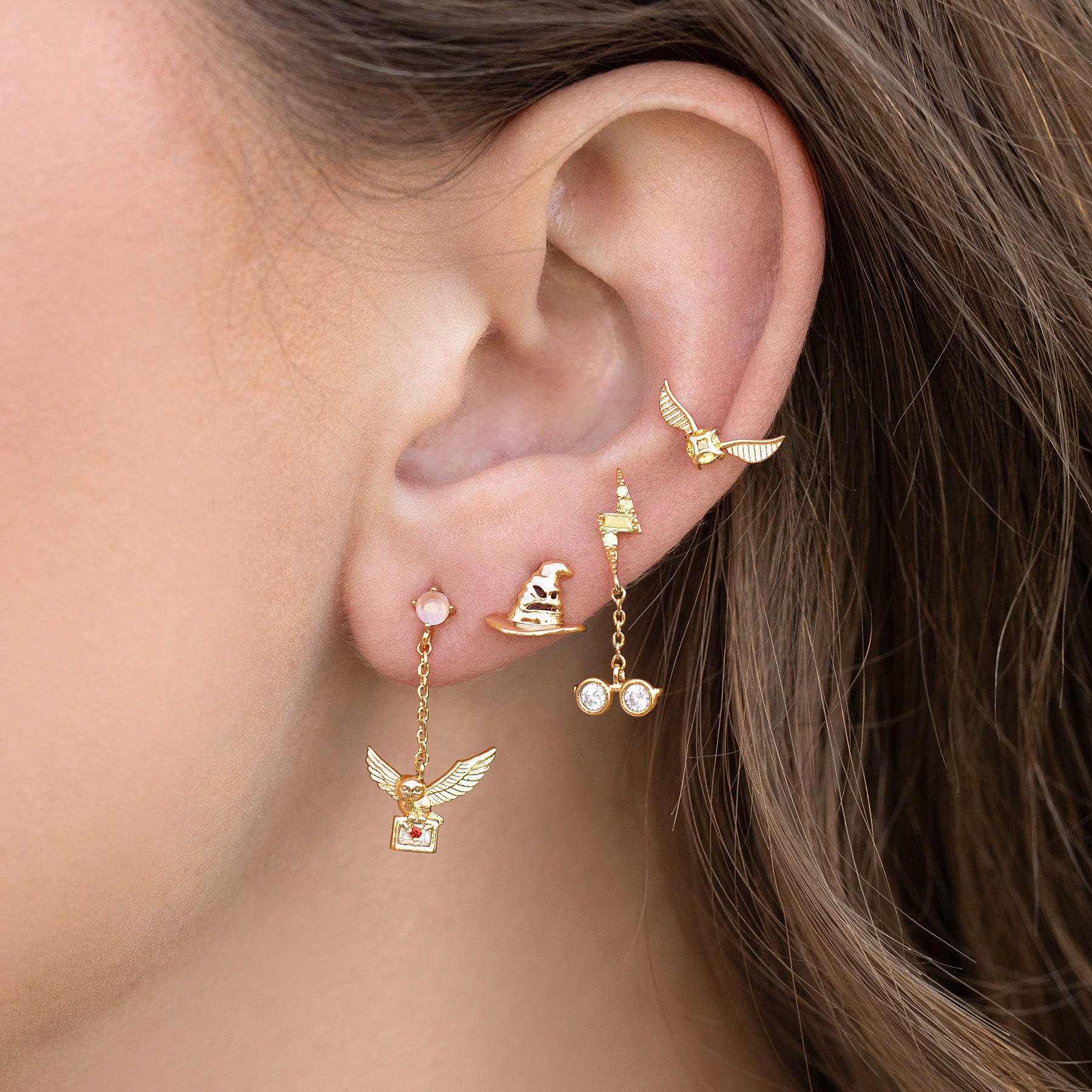 Close-up of an ear with gold earrings featuring the golden snitch and small gemstones.