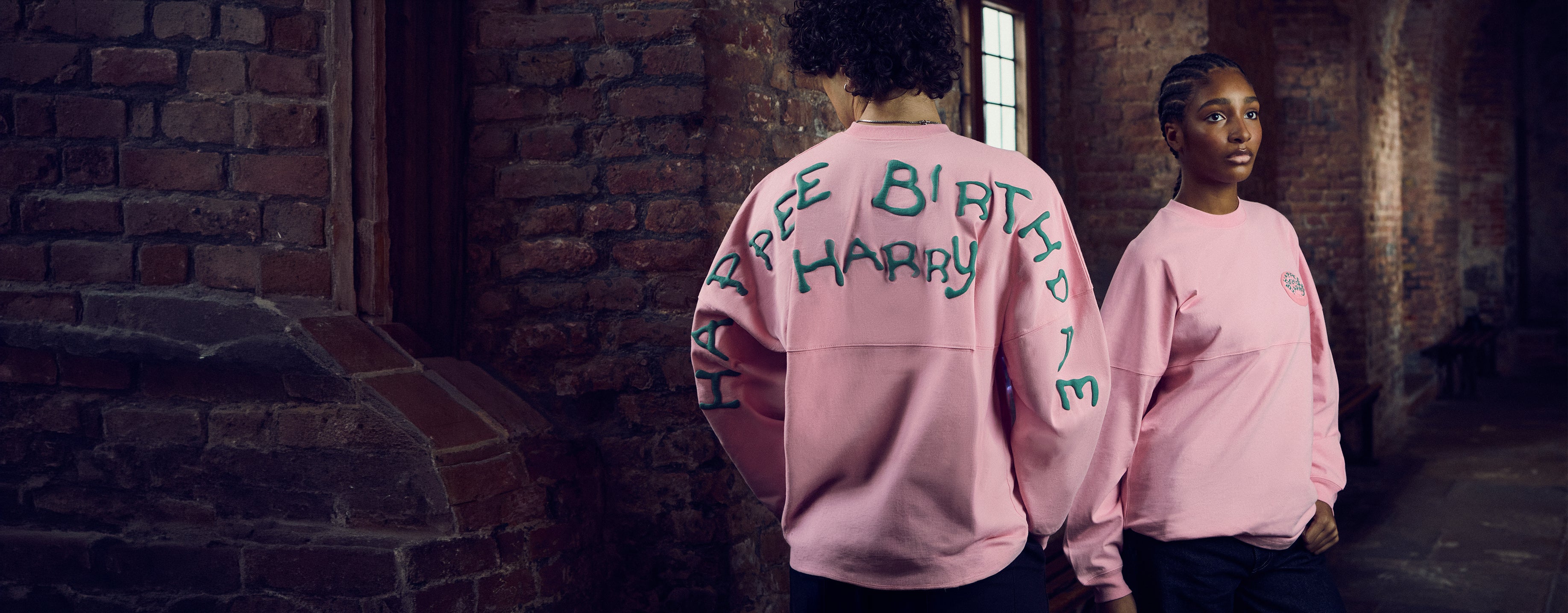 Two people wearing pink Happy Birthday Harry sweatshirts in a brick-walled corridor.