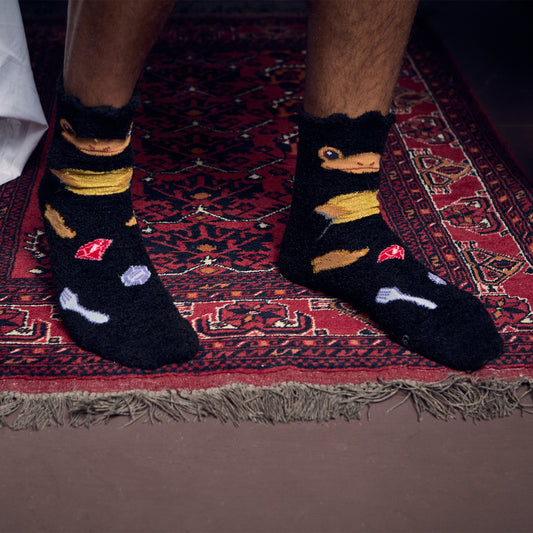 A person wearing black Niffler novelty socks with cartoon Niffler faces and small colorful gem-like motifs, standing on a red patterned rug.