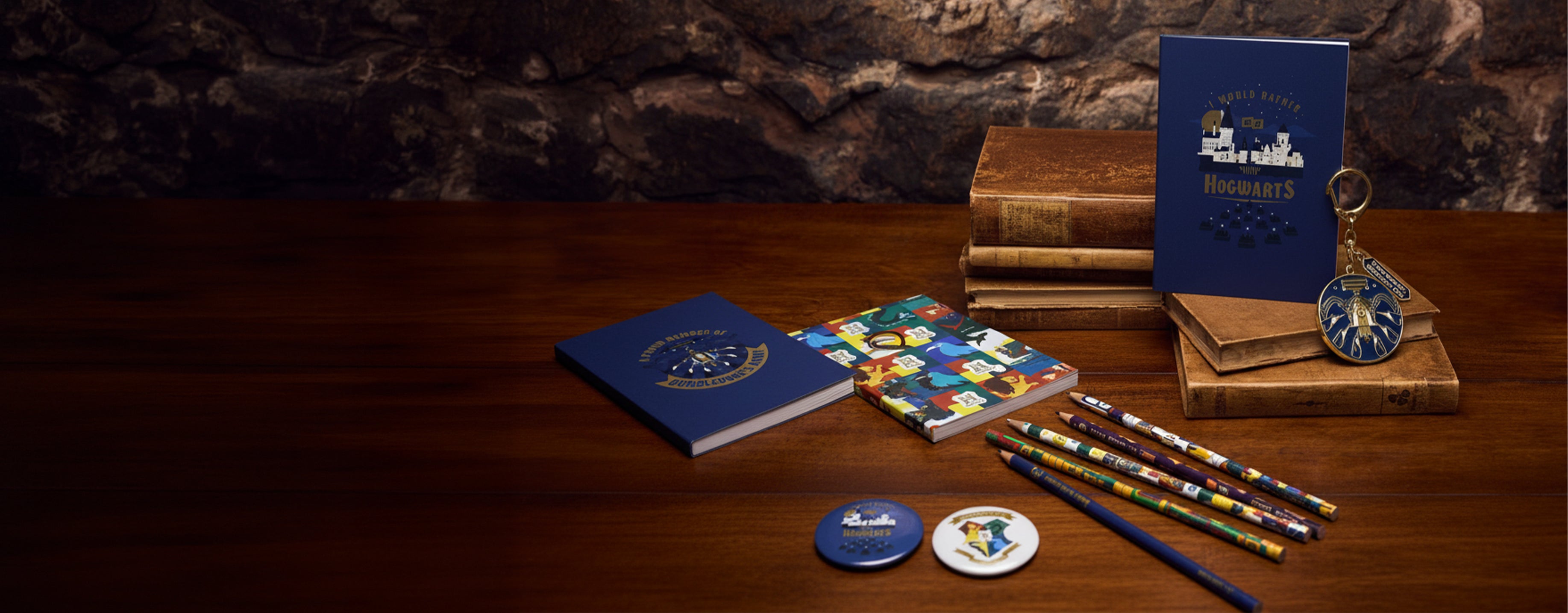 A collection of blue and multicolor stationery items arranged on a dark wooden table against a rustic stone wall background. The set includes three notebooks, a blue keyring, two small round pins, and several pencils.