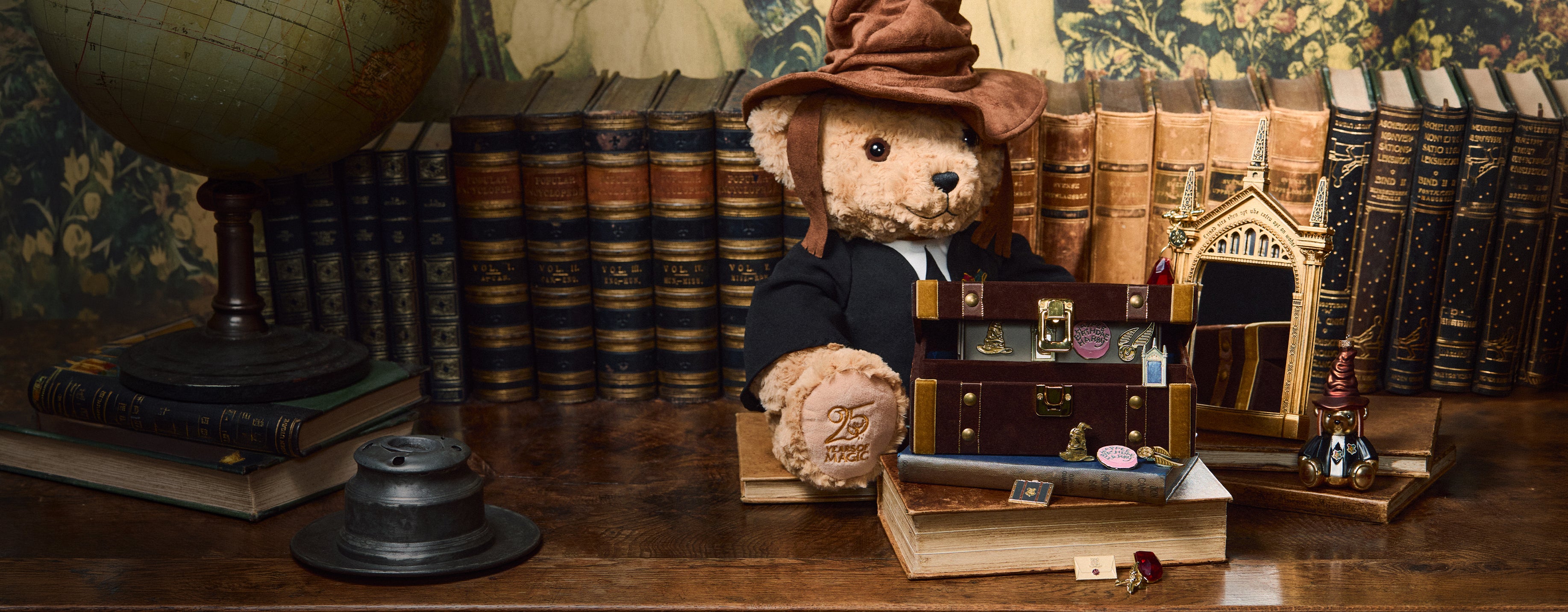 Teddy bear in a Hogwarts robe wearing the Sorting Hat, surrounded by books and a mirror on a wooden table.