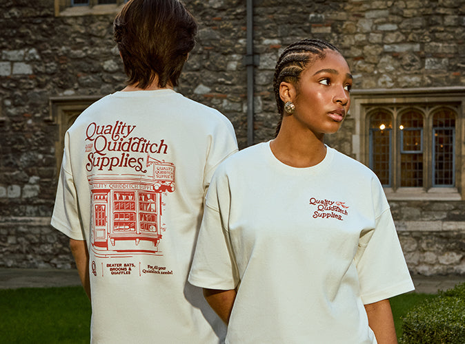 Two people wearing 'Quality Quidditch Supplies' t-shirts in front of a stone building.