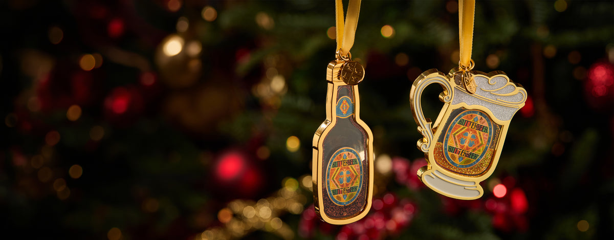 Two decorative hanging ornaments shaped like a Butterbeer bottle and a Butterbeer stein, both with gold trim and colourful printed graphics, suspended in front of a blurred Christmas tree background with red ornaments and warm lights.